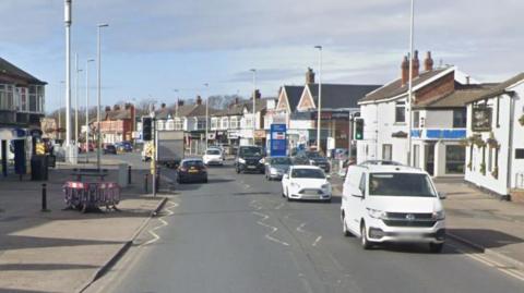 Google streetview of vehicles driving along Whitegate drive with shops and properties on either side.