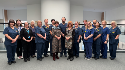 A large gathering of men and women in dark blue hospital uniforms standing inside a white-walled building with a grey carpet, in front of railings 