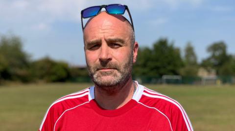 Bearded man with sunglasses on his head, wearing a red sports t-shirt, standing on a grassy field with trees and goal posts behind him