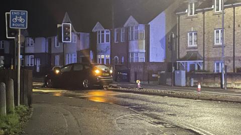 A black car on the central reservation of a residential road having just hit some traffic lights. There are terraced houses lining the street and orange and white cones. 