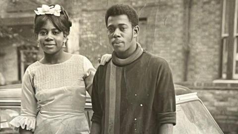 A black and white image of a man and a woman standing in front of a white car. The man is wearing a dark jumper with a stripe on the right hand side. The woman has her hand on his shoulder and is wearing a light dress and white headpiece