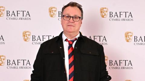 Russell T Davies attends the 2024 BAFTA Cymru awards wearing a black jacket and a red and blue striped tie. He is wearing glasses.