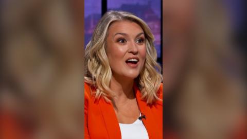 Dragon Sara Davies has shoulder length wavy blond hair and wears an orange blazer over a white top