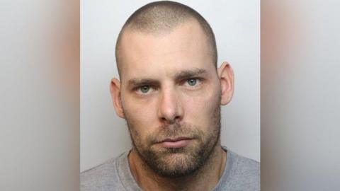 A mugshot of Damien Bendall. He has short, shaved hair and a lightly grown beard and moustache. He has green eyes and is wearing a grey T-shirt.