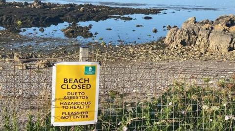 An empty Mount Batten beach near Plymouth which has been closed following the discovery of asbestos. There is a yellow warning signing saying "BEACH CLOSED". The beach is stoney with lots of brown seaweed on it. The water is dark blue coloured with bright blue skies above it. 
