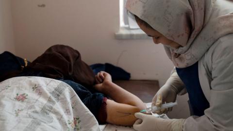 A side view of nurse wearing a headscarf attending to a female patient on a hospital bed. The patient's face is covered and the nurse is turned away from the camera.