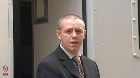 Nicky has a shaved head. He is wearing a grey suit jacket and tie.