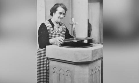 In a black and white photograph Mabel Wulff stands at a stone font in the English Church in Hamburg. Dressed in a cheque pinafore with her dark hair pinned back, she's working in the church.