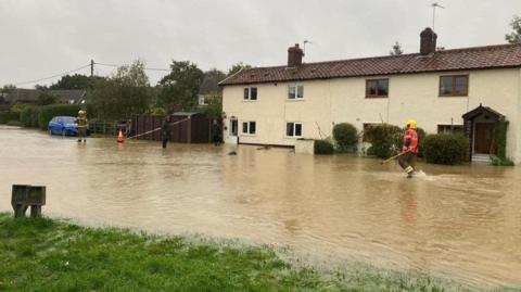 A row of cream-coloured houses looking out over a wide body of water, which goes up to the front doors. A firefighter wades through the water while another firefighter is on the left.
