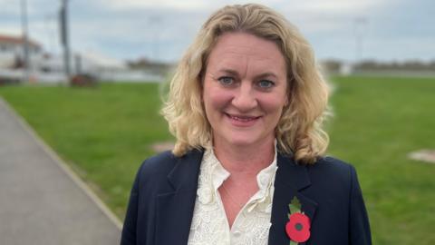 Louise McKinlay stands on a pathway in front of a section of green grass. She has shoulder length blonde hair that is slightly curled. She wears a navy suit jacket with a poppy on one side of her chest and a cream blouse underneath.