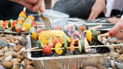 A single-use barbecue is sitting on top of pebbles. There is a corn on the cob and skewers with vegetables on them on top of the barbecue.