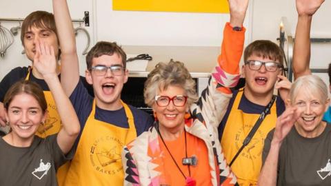 A group of people including trainees, staff members and Prue Leith smile and wave at the camera. Most are wearing Yellow Submarine t-shirts or aprons.