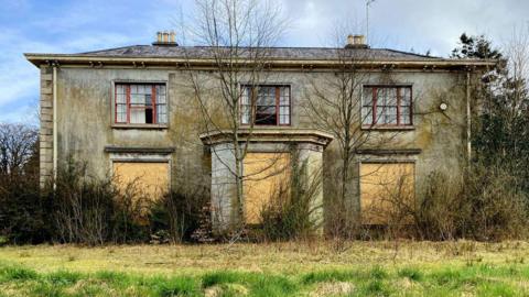 Crevenagh House. It is a two storey grey building with broken windows on the top floor and boarded up windows at the bottom. There are overgrown plants at the front of the building.