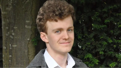 Sam Carling smiles and looks directly at the camera. He is photographed outside in front of green foliage and is wearing a white shirt and grey jacket. 