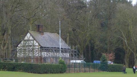 A black and white timber building covered in scaffolding is surrounded by hedges and trees in a park.