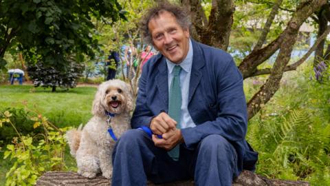 A smiling Monty Don sitting on a log with a dog in the garden at Battersea