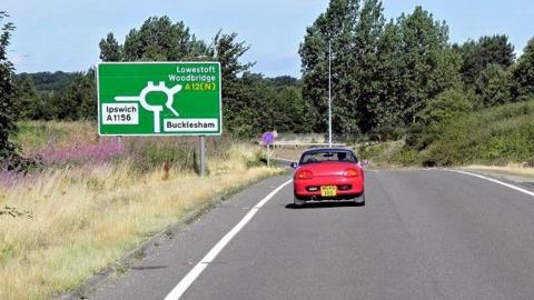 A slip road from the A14 approaching the Seven Hills junction of the A12. A red car is driving by a green road sign.