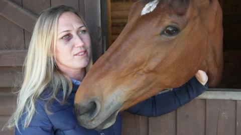 Becci stood next to a stable door with Barney - a brown horse - sticking his head out. Becci, who has long blonde hair and is wearing a blue coat, has a black eye from when she was knocked off Barney. 