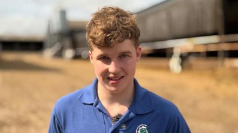 A young man is on a farm. There are sheds in the background.