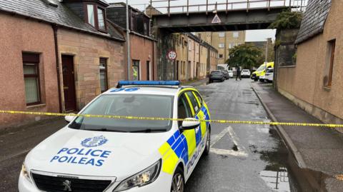 A police car is parked in a street in Merkinch in Inverness. A piece of police tape is stretched across the street and over the front of the car. The street is lined by brown walled buildings and up the street is a railway bridge.