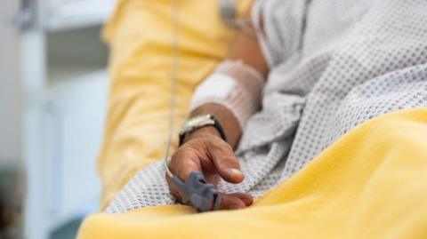 A man's hand in a hospital bed with a pulse monitor 