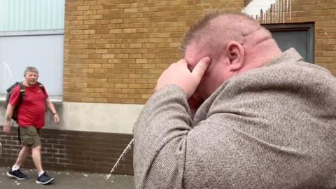 Side profile of Bernard McDonagh in a street. He holds his hand to his face covering his eyes. He has fine short hair and wears a brown tweed suit jacket. A man can be seen in the background wearing a red t shirt, cargo shorts and black trainers. 