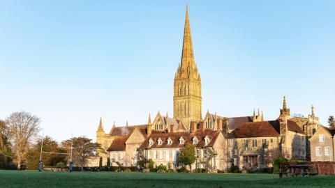 A cathedral spire in the background of several historic stone buildings and a playing field with rugby goal posts at one end