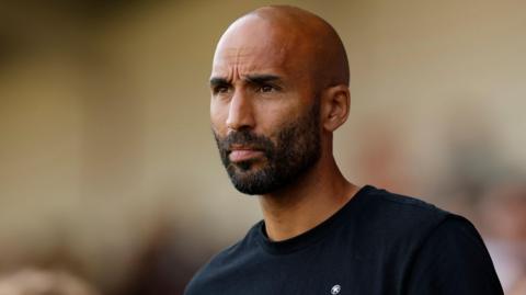 Walsall head coach Lee Grant watches his side from the touchline. He is wearing a dark sweatshirt with Walsall written in white on front.