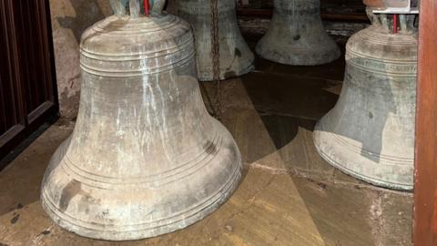 Sitting on the floor are metal bells one of which is half a metre in size. There are four in the picture but there are a total of 5 bells.