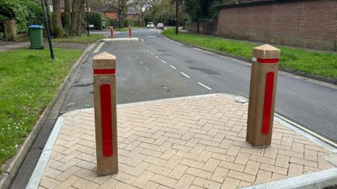 A shot of a road, flanked by strips of grass and trees on either side. In the foreground are two wooden bollards atop a raised paved platform. Another platform and bollards can be seen a little further down the road. 