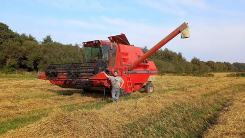 A man wearing muddied blue jeans and a grey T-shirt standing in a field next to a large red threshing machine. The crops have just been cut and are yellow and green.