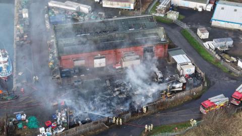 Aerial view of the smouldering aftermath of a fire at a yard next to a harbout. Two fire engines and several firefighters can be seen in the image.