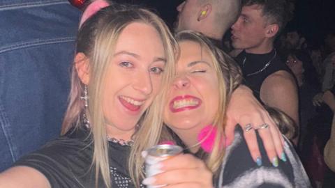 Abi takes a selfie using a digital camera. She smiles widely at the camera. She has long blonde hair and wears a black crop top with a blue jumper tied round her waist. She has her arm around another woman who has long blonde hair and smiles widely at the camera. She wears a black T-shirt and holds a can of drink.
