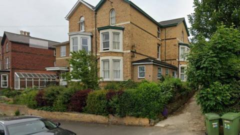 A large brick house surrounded by privet hedge and shrubs with large bay windows.