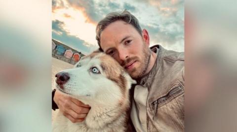 Ben Gladman, wearing a light brown jacket, pictured with his husky/malamute dog called Dobby. He is on a beach and there are beach huts behind him.