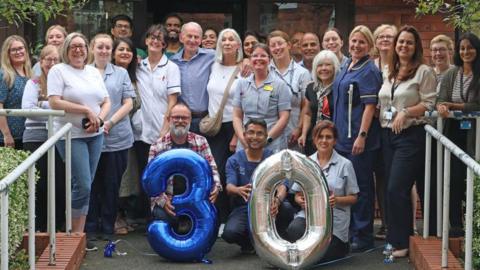 A group of about 25 people stand at the entrance to a building, posing for a photograph, with three kneeling down at the front holding balloon numbers three and zero.