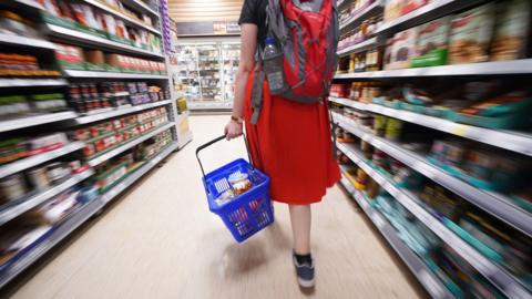 A girl in a red dress is carrying a blue shopping basket as she walks along the aisle of a supermarket. 