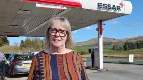 Judith stood outside her fuel garage on a sunny day. She looks at the camera with a stern expression. She has short grey hair and wears brown glasses, a multi coloured top and gold necklace.