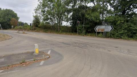 A T-junction on a road with trees in the background and road signs and traffic lights in the distance.