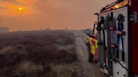 A four wheel drive Land Rover fire engine is at the top of a path, with firefighters silhouetted against an orange sky surrounded by smoke, in the background.