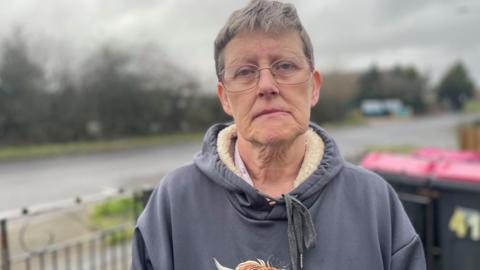 A women with short grey hair and glasses stands alongside a wide road looking dejected