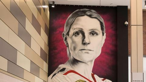 A mural of a woman wearing an England football top. There is a column to the right and a wall on the left which is patterned in grey and light brown rectangular shapes. 