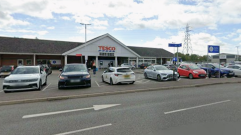 A supermarket car park, with a row of cars in front of the main entrance to the store. There is an electricity pylon in the distance and directional arrows on the road in front of the vehicles.