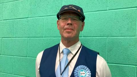 A man in a white shirt with blue waistcoat and blue cap and light blue tie and light blue Reform UK rosette standing against a green wall