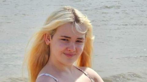 A photo of Phoebe Macias, who has long, bleached blonde hair and wears a strappy top as she looks at the camera, with the sea behind her.