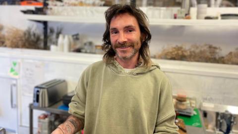 Nathan who has shoulder-length dark hair and a short beard, smiles at the camera behind the counter in his cafe. He is wearing a light green hoodie. 