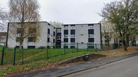 A streetview photo of identical three-storey blocks of flats in grey render and dark cladding in between. In front, there is green security fencing separating the grass around the flats from the road in front.