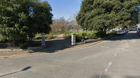 Entrance to Devonport Park. Two pillars either side of the park where people can walk through. There is a junction on the road outside with two big trees either side of the walkway. 