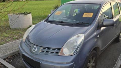 A grey Nissan Note car, which is parked in a car parking bay. Next to it is green grass and planters. On the car's window are two yellow stickers that read "seized" in black writing.