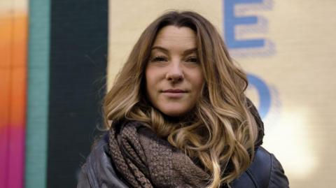 A woman with long brown hair stands in front of a colourful building. She is wearing a dark jacket and a chunky brown scarf wrapped around her neck.